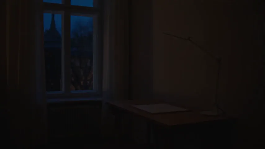 Prueba de Seedance 2.5 con el prompt de la lámpara de escritorio: un diseñador ajusta una lámpara blanca sobre una mesa vacía y, después, el clip salta a un primer plano del brazo de la lámpara delante de un boceto de un edificio clavado en la pared, con una mano desenfocada en primer término.