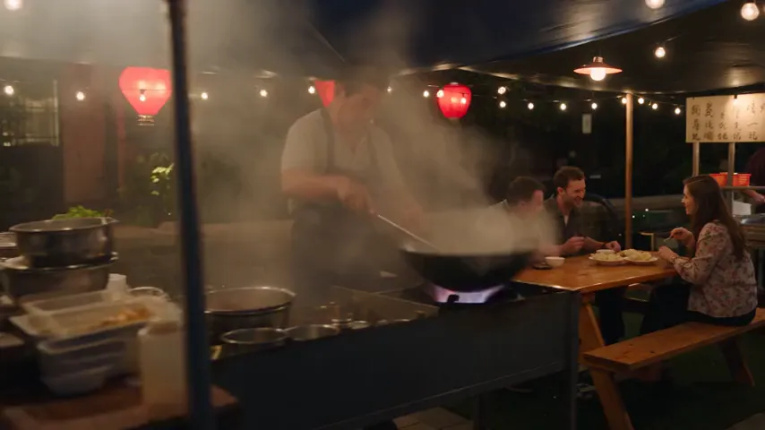 Seedance 2.5 test render of the night market prompt: a cook at a steaming wok next to diners at a wooden table, the diners laughing under string lights, and a large handwritten sign with made-up characters beside the stalls.